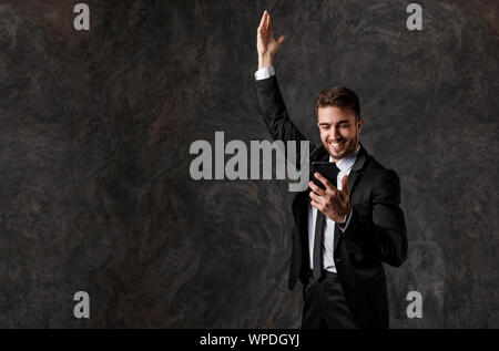 Un giovane imprenditore barbuto in una tuta guarda al telefono e gioisce. uomo di successo ha visto buone notizie sul telefono, gioia e glee dalla chiusura di una trattativa. Foto Stock