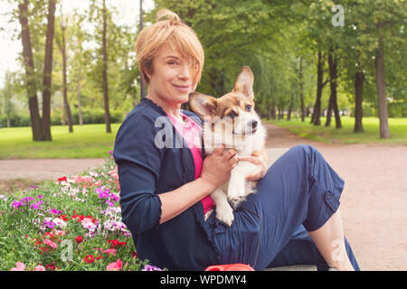 Felice anziani donna seduta con il cane sul banco in fiore d'estate il parco Foto Stock