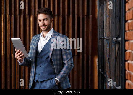 Giovane imprenditore di successo con la barba in un lussuoso blu tuta controllati. L'uomo lavora su un tablet in un loft ufficio. Foto Stock