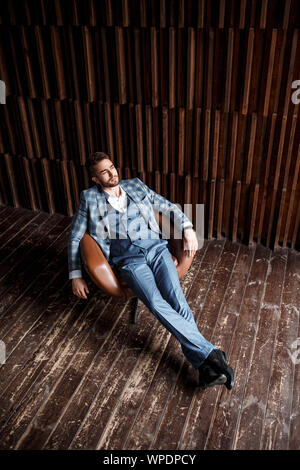 Giovane imprenditore di successo in una tuta blu in una gabbia è seduto in una sedia in pelle. giovane uomo con la barba sorridente Foto Stock