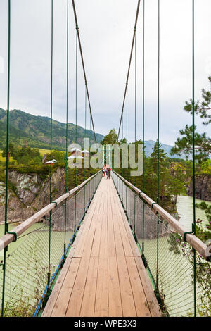 Vista della strada attraverso una sospensione in legno ponte con imbracature in ferro che conduce ad un'isola rocciosa con una chiesa in un verde fiume di montagna. Il concetto Foto Stock