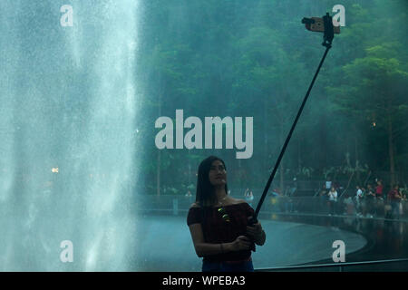 Donna asiatica usando un bastone selfie per scattare foto in piedi di fronte ad un vortice di pioggia al gioiello in aeroporto Changi Foto Stock