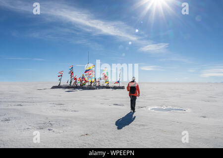 Giovane uomo che cammina per le bandiere internazionali provenienti da diversi paesi a Uyuni distesa di sale in giornata soleggiata con raggi solari, Bolivia, Sud America travel voc Foto Stock