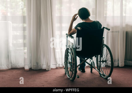 Premuto triste donna in sedia a rotelle usurati guardando fuori dalla finestra e pensare, lonely disabile adulto persona caucasici in ambienti interni Foto Stock