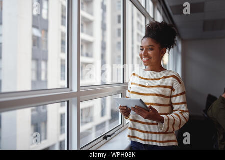 Riflessivo sorridente bella giovane americano africano imprenditrice con tavoletta digitale in piedi alla finestra di ufficio che guarda lontano Foto Stock