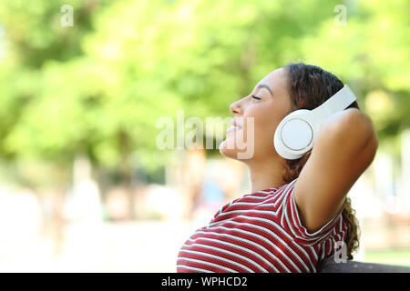Vista laterale verticale di un felice razza mista donna ascoltando musica in appoggio seduta su una panchina nel parco Foto Stock