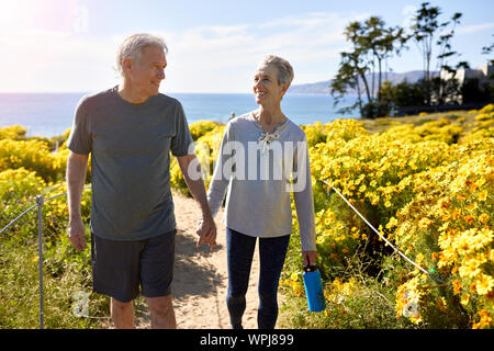 Sorridente coppia senior tenendo le mani mentre si cammina sul sentiero in mezzo a piante sulla scogliera dal mare contro il cielo durante la giornata di sole Foto Stock