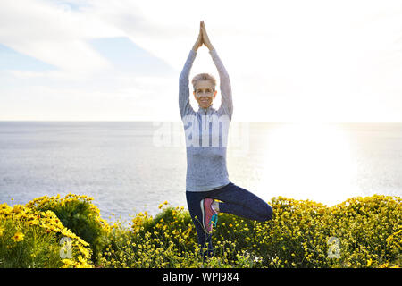 Ritratto di sorridente donna senior facendo la posizione dell'albero mentre si esercita nel mezzo di piante a mare contro il cielo durante la giornata di sole Foto Stock