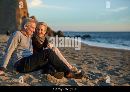 Coppia senior usando il telefono mentre è seduto sulla sabbia in spiaggia durante il tramonto Foto Stock