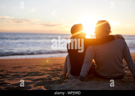 Vista posteriore della coppia senior di parlare mentre è seduto sulla sabbia in spiaggia durante il tramonto Foto Stock