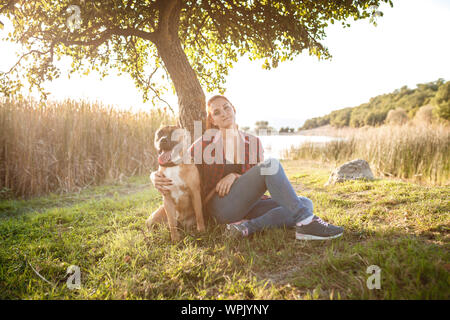 Jouful giovane ragazza che accarezzano il loro cane, indossare abbigliamento sportivo, godendo il loro tempo e la loro vacanza nella soleggiata park Foto Stock