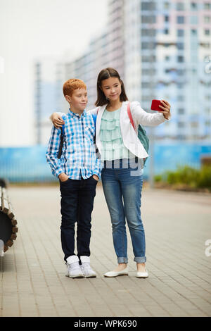 Giovane ragazza asiatica rendendo selfie ritratto sul telefono mobile insieme con il suo amico mentre sono in piedi e abbracciando nella città all'aperto Foto Stock