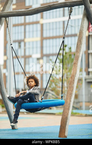 Ragazzo africano con i capelli ricci e in abbigliamento elegante guardando la fotocamera durante l'oscillazione sull'altalena all'aperto Foto Stock