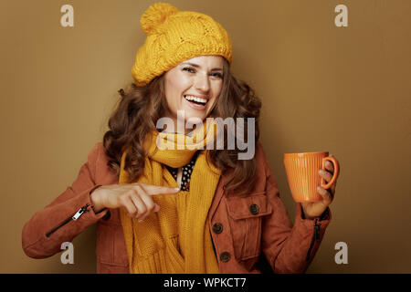 Ciao autunno. Sorridendo donna moderna in giallo berretto e sciarpa puntando alla tazza di latte macchiato di zucca contro lo sfondo di bronzo. Foto Stock