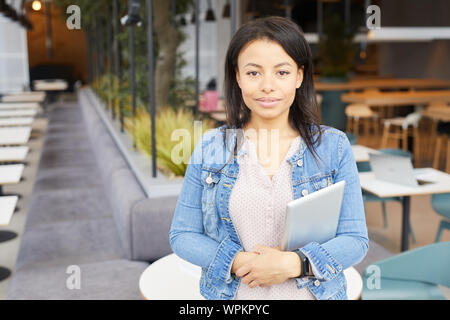 Ritratto di giovane donna bellissima con scuri capelli lunghi in abbigliamento casual azienda digitale compressa e guardando la telecamera mentre in piedi presso il ristorante Foto Stock
