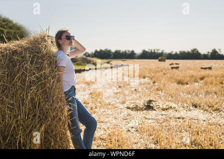 Giovane donna con capelli castani in T-shirt bianco e blu jeans pone dal pagliaio su un campo dopo il raccolto. Calde luci del tramonto Foto Stock