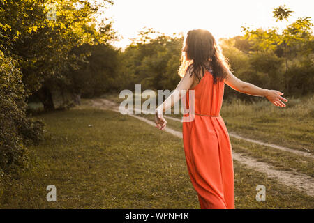 Bella ragazza giovane con scuri capelli ricci in arancio brillante abito di passeggiate in la distanza attraverso la foresta o campo al tramonto. Umore romantico. Solitudine Foto Stock