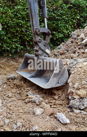 Scavo di un concreto passo carraio al fianco di una siepe; metallo benna di scavo si appoggia su un gran mucchio di rotture di calcestruzzo e mattoni Foto Stock
