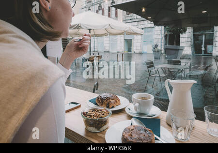 Vista posteriore della sola donna mangiare delizioso yogurt con muesli di bere il caffè con croissant e torte al mattino presto in francese cafe con si affacciano alla piazza centrale terrazza Foto Stock