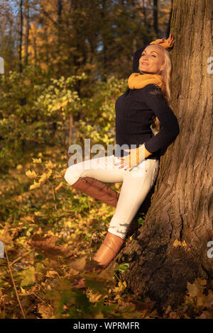 La ragazza si appoggia su un albero e viene fotografata al tramonto in abiti da cowboy, stivali marroni gamba su un albero faccia a fianco Foto Stock