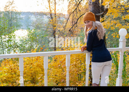 Una ragazza nella foresta autunnale si trova sul bordo di un ponte bianco e guarda il fiume con foglie gialle brillanti Foto Stock