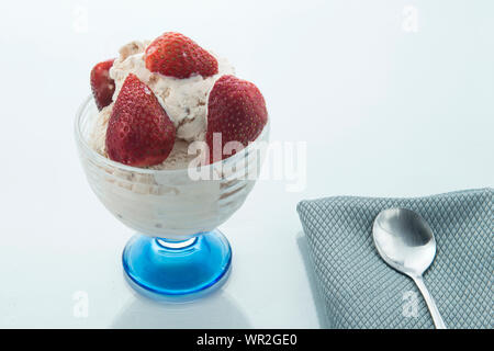Le fragole in un bicchiere di vetro e più fragole su una piastra blu su bianco e superficie lucida e una forcella sul lato, sfondo bianco. Foto Stock