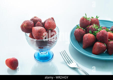 Le fragole in un bicchiere di vetro e più fragole su una piastra blu su bianco e superficie lucida e una forcella sul lato, sfondo bianco. Foto Stock