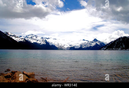 Vibrant Blue Lago Garibaldi con ghiacciaio nevoso monti all'orizzonte e di un bianco luminoso cielo nuvoloso, su un sentiero escursionistico in Garibaldi Provincial Park Foto Stock