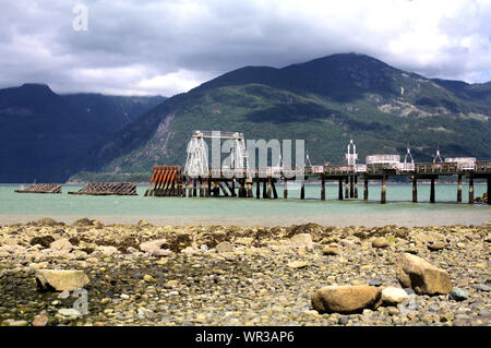 Il vecchio molo Porteau Cove Parco Provinciale in un giorno nuvoloso, con la spiaggia rocciosa, turchese-fiordo di verde e le montagne sullo sfondo Foto Stock