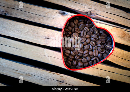 I chicchi di caffè in un cuore-stagno sagomati disposti su un sfondo di legno. Foto Stock