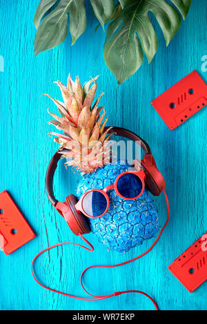 Tanga carattere di ananas in rosso gli occhiali da sole e auricolari ascolta la musica. Rustico in legno di colore blu con sfondo color corallo nastri audio e exoti Foto Stock