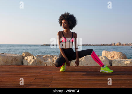 Felice giovane donna africana di stretching e riscaldamento prima di un allenamento all'aperto. Il mare Mediterraneo in background Foto Stock
