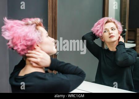 Bella acconciatura di giovane donna guardando nello specchio dopo la morte dei capelli e rendendo mette in evidenza in un salone di bellezza. Vicino a breve taglio di capelli rosa Foto Stock