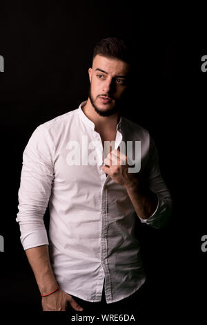 Moda ritratto di Giovane Uomo in camicia bianca si pone al di sopra della parete nera con contrasto ombre e ha perso il contatto con gli occhi. Foto Stock