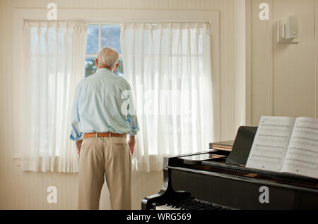 Senior uomo guardando fuori dalla finestra accanto ad un pianoforte. Foto Stock