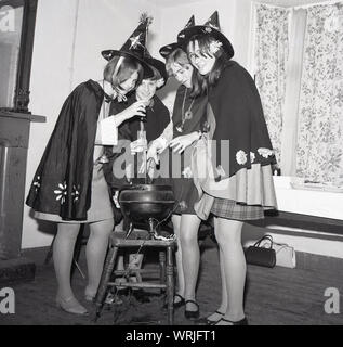 1967, storico, quattro ragazze adolescenti vestiti con costumi delle streghe - nero cappotti e cappelli di punta - in una stanza intorno a una ghisa pentola o paiolo di rame appollaiato su uno sgabello, la preparazione di un magico di vestirsi o "streghe brew' Inghilterra, Regno Unito. Foto Stock