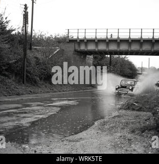 1967, storica strada allagata, auto d'epoca andare sotto un ponte attraverso una grande piscina di acqua su una strada dove gli scarichi hanno traboccato. Risborough, Inghilterra, Regno Unito. Foto Stock