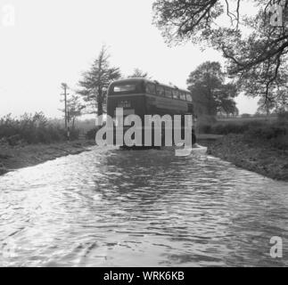 1967, storico, una strada allagata, un double-decker bus diretto a Thame, Bucks, passando attraverso una grande piscina di acqua sulla strada di un paese dove gli scarichi hanno traboccato. Foto Stock