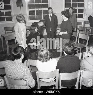 Anni sessanta, storico, filodrammatica, persone in un dramma gruppo provare un gioco in village hall, guardato da altri giocatori, Inghilterra, Regno Unito. Foto Stock