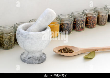 Stone mortaio con pestello, grande cucchiaio di legno con pepe macinato e vasetti di vetro con diverse spezie su una cucina bianca tabella. Utensili moderni. Foto Stock