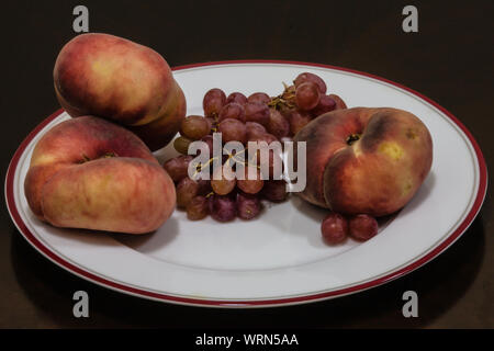 Pesche piatte (Prunus persica var. platycarpa) e uva rossa su una piastra bianca Foto Stock