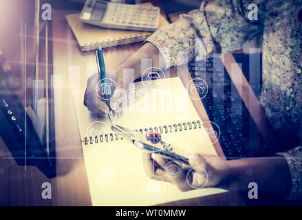 Le mani della donna che lavorano sulla scrivania in ufficio che una mano che tiene una smart phone e scrivere su notepad con grafica virtuale diagramma icona. Foto Stock