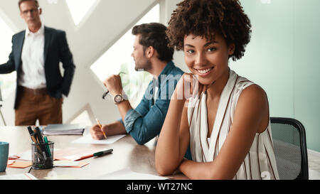 Sono felice di lavorare qui giovani afro american donna sta guardando la telecamera e sorridere mentre il suo boss in piedi vicino a lavagna e discutere di qualcosa con il team. Il lavoro di squadra. Sale riunioni Foto Stock