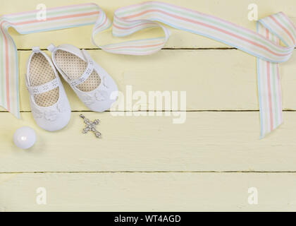 Battesimo con sfondo bianco,scarpe nastro, candela e cristallo pendente a croce su dipinte di giallo chiaro dello sfondo di legno - vista superiore foto Foto Stock