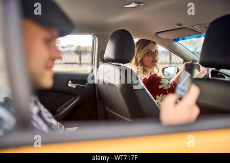 Foto del giovane nel cappuccio con il telefono in mano seduto nel sedile posteriore, le donne la guida nel pomeriggio Foto Stock