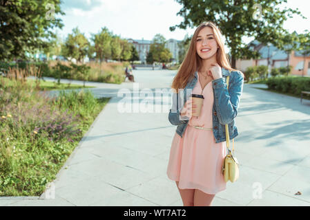 Bella ragazza abito rosa felice ridere sorridere gioiosa, tenendo tazza di tè del caffè mano, estate city park, sulla luminosa giornata di sole. Emozioni delizia e Foto Stock