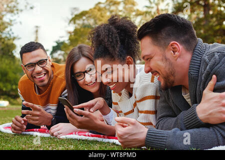 Sorridente razza mista gruppo di amici giacente insieme su erba verde utilizzando il telefono cellulare nel parco - amici avente un picnic Foto Stock