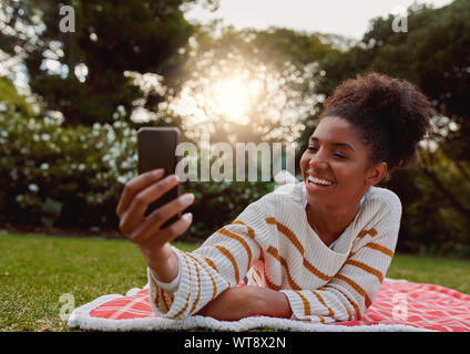 Felice giovane donna africana giacente su erba verde tenendo selfie attraverso il telefono cellulare sulla giornata di sole Foto Stock