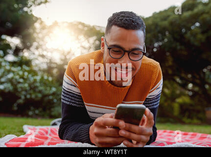 Ritratto di un africano sorridente giovane studente nero da indossare occhiali relax nel parco utilizzando smart phone - studente di college utilizzando mobile in posizione di parcheggio Foto Stock