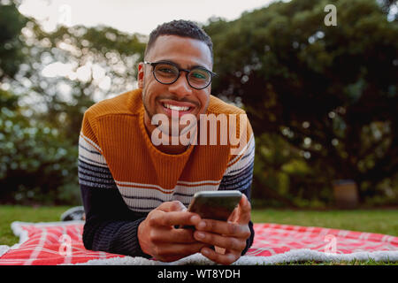 Ritratto di un sorridente americano africano giovane uomo disteso sulla coperta nel parco tenendo il telefono cellulare in mano guardando sorridente della fotocamera
